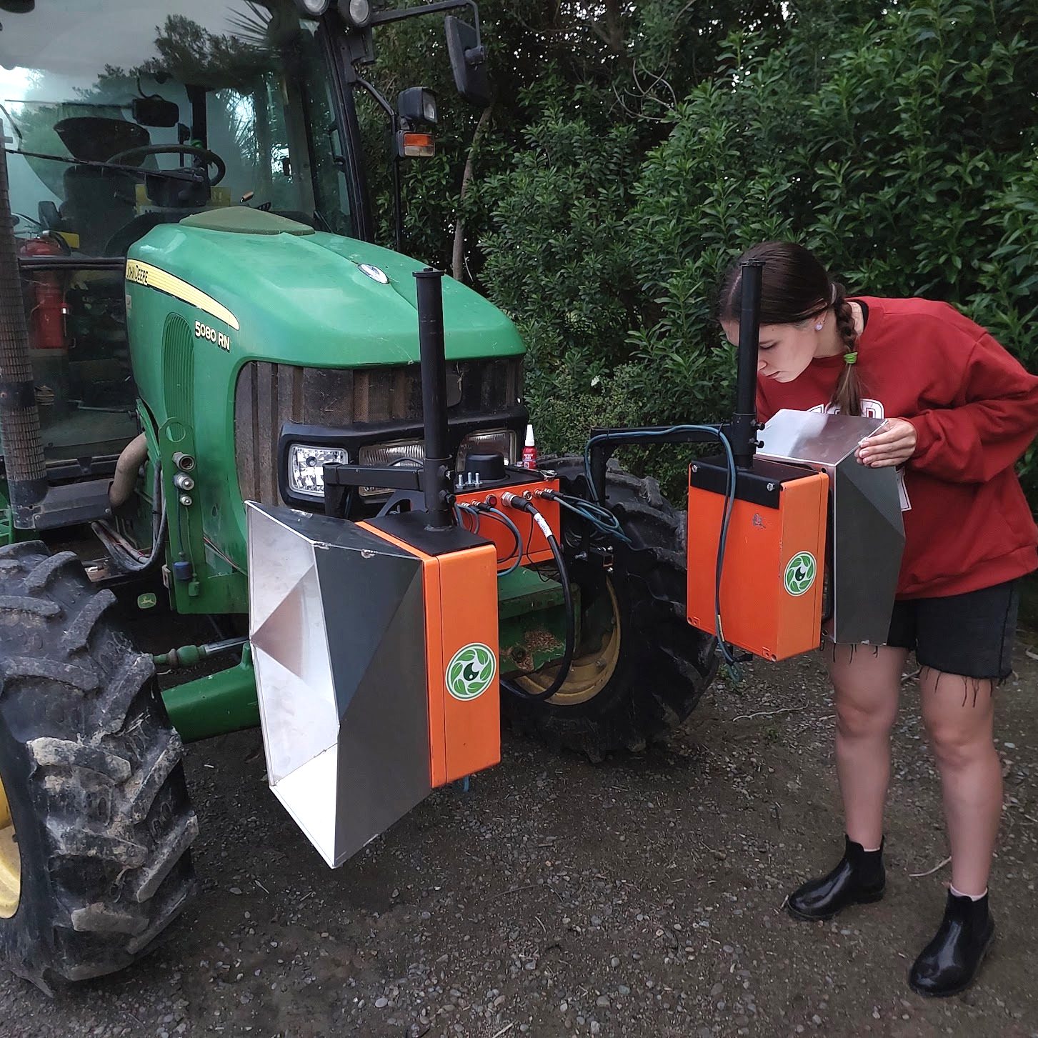 A green John Deere 5080 RN tractor fitted with two orange Cropsy scanner units and white calibration targets, a team member alongside taking notes
