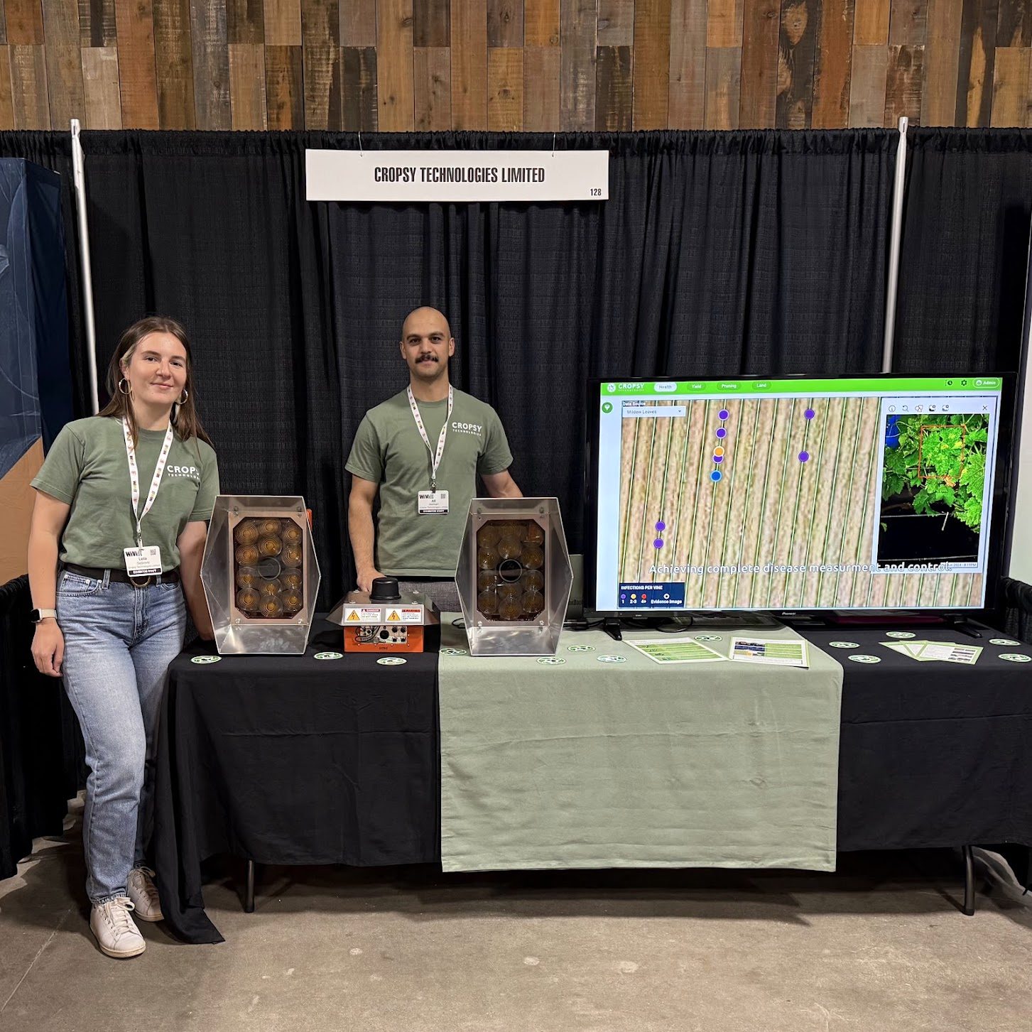 Two Cropsy team members at an exhibition booth, two scanner units on the table and vineyard maps on the screen behind
