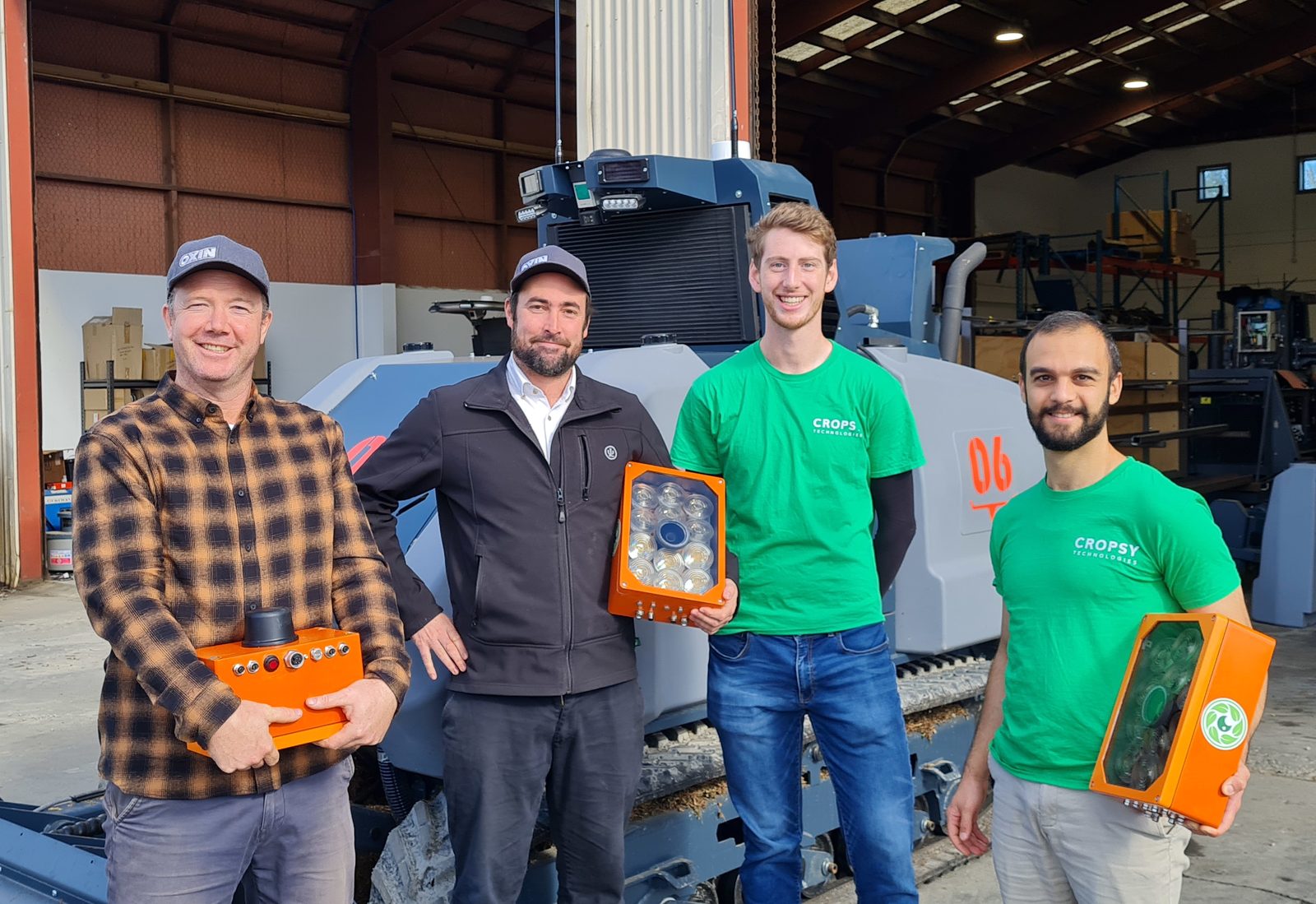 Four people in a machinery shed, each holding an orange Cropsy scanner module, in front of an OXIN autonomous vineyard machine